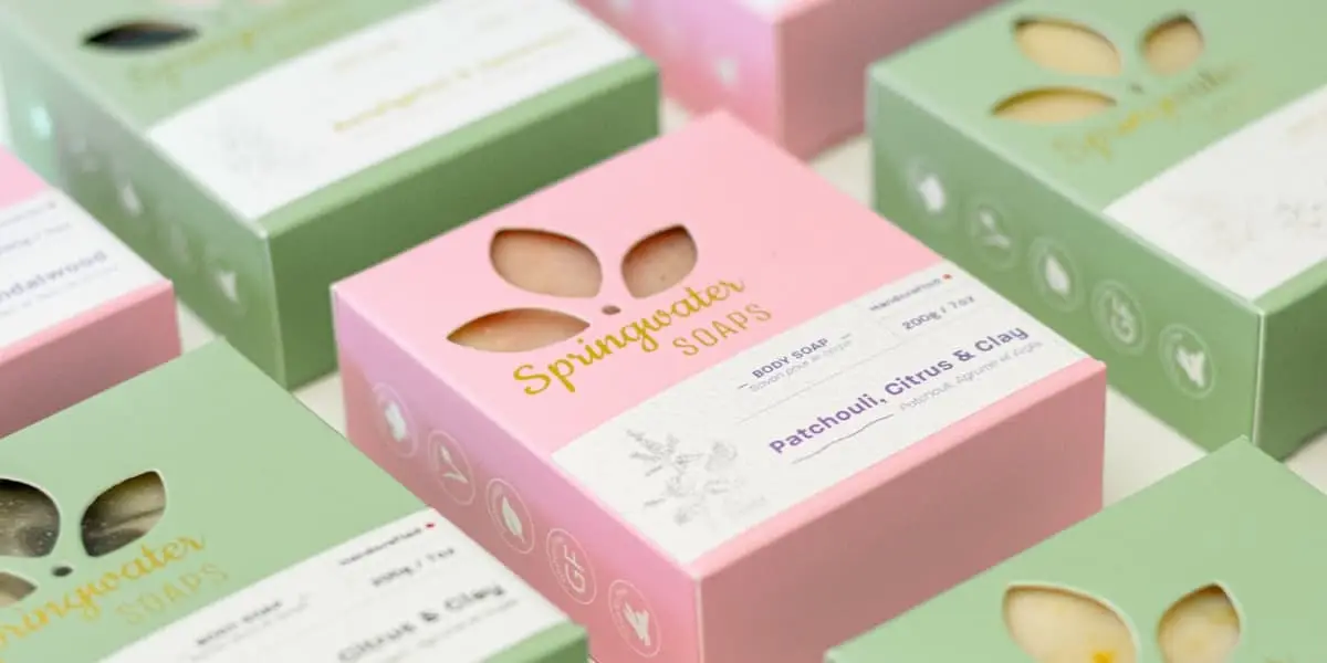 Cajas de jabón artesanal en rosa y verde alineadas sobre una superficie clara